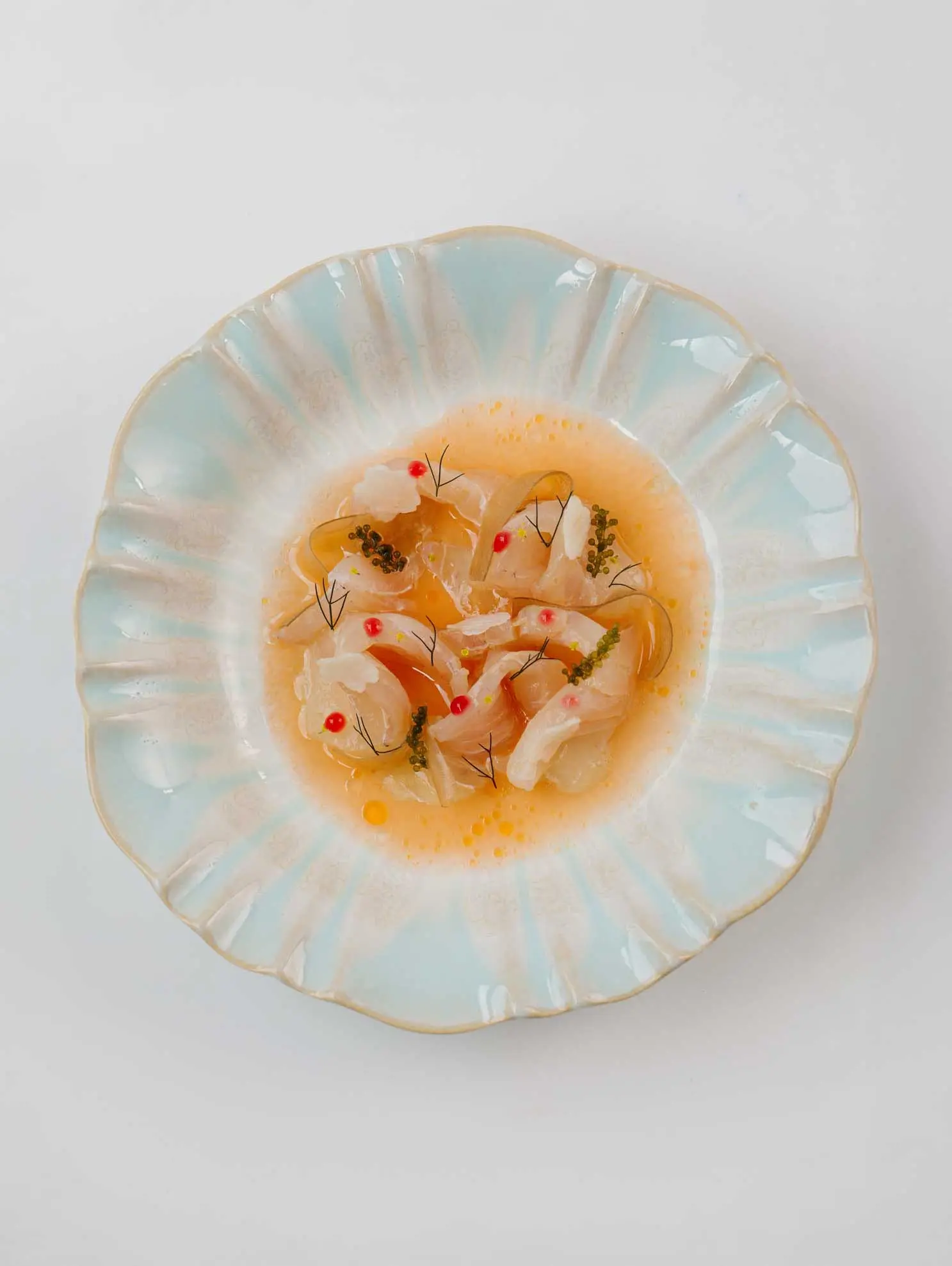 Slices of raw white fish in a citrus broth with sea grapes and redcurrants, in a pale blue scalloped bowl