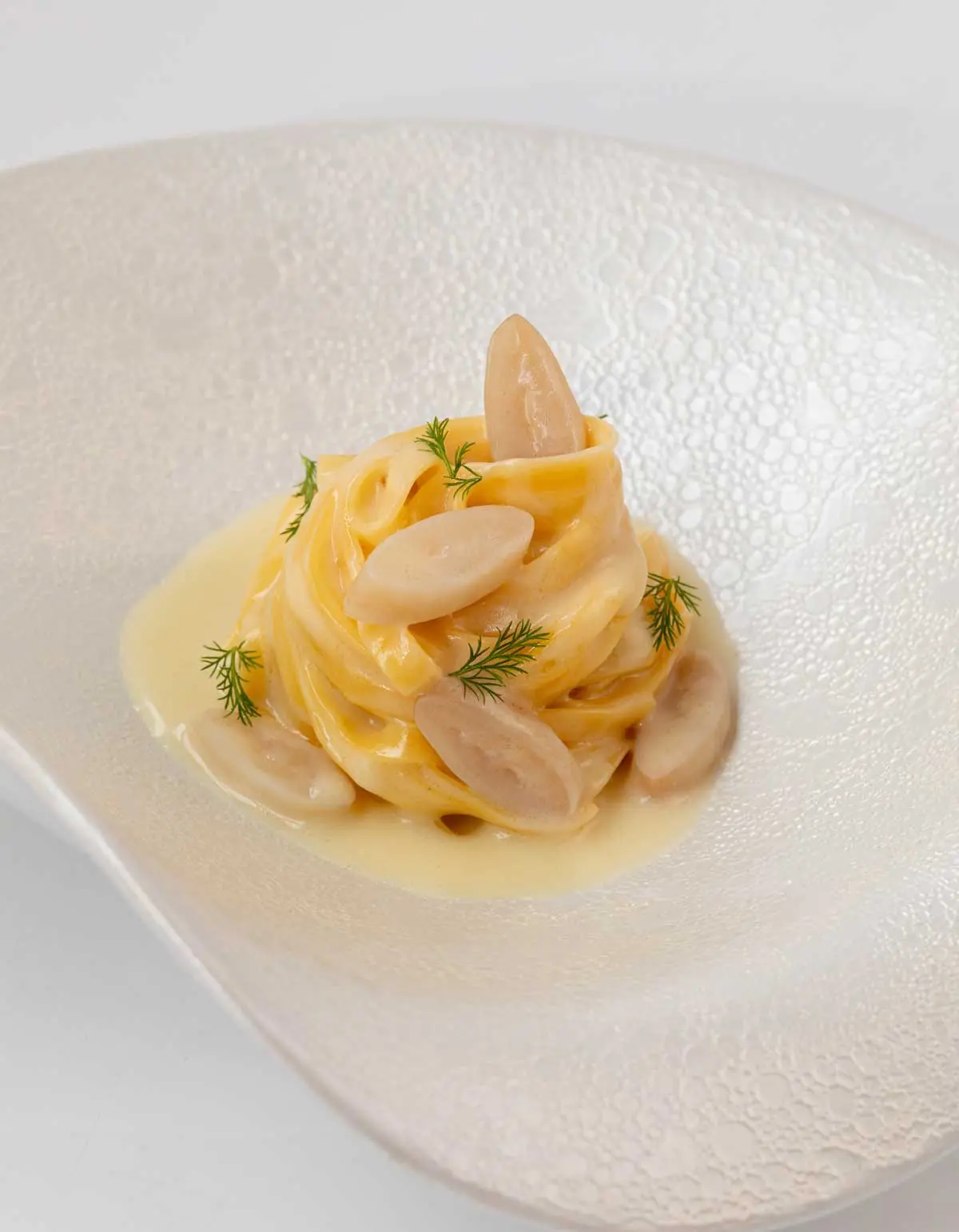 Nest of tagliolini in glossy butter sauce with pale sliced bulbs and dill, in a textured white bowl