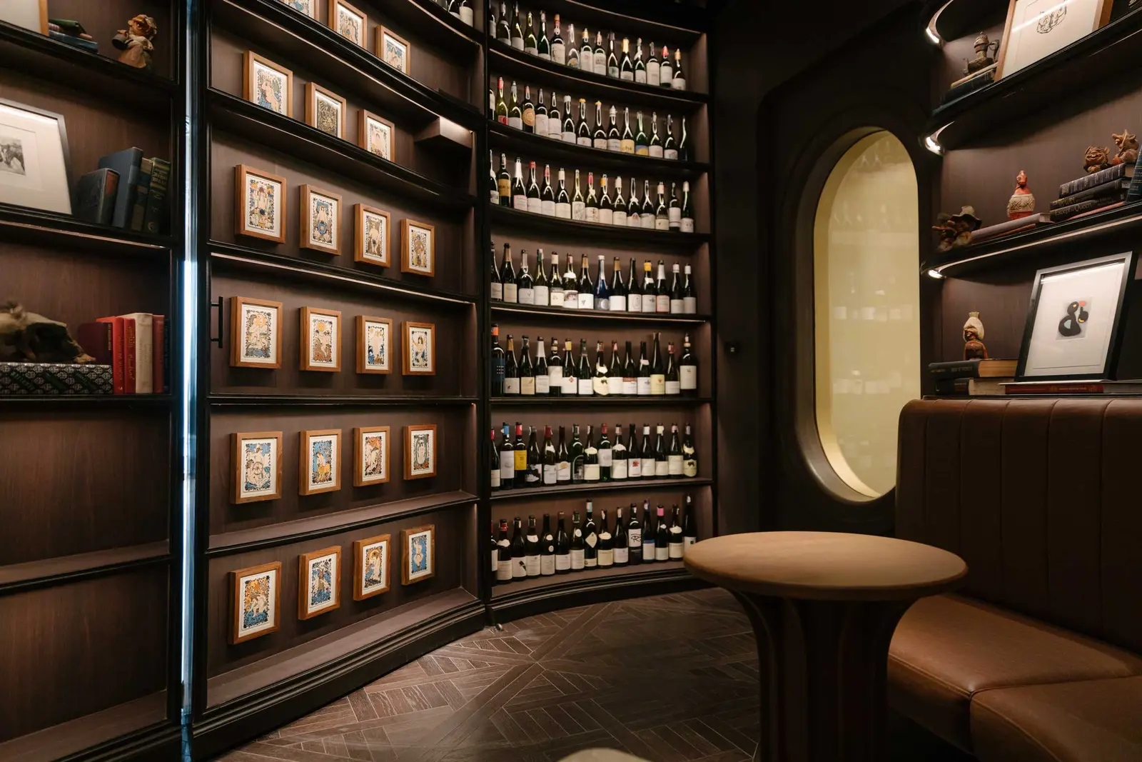 Curved dark-wood wine room with bottle-lined shelves, framed illustrations and a leather banquette