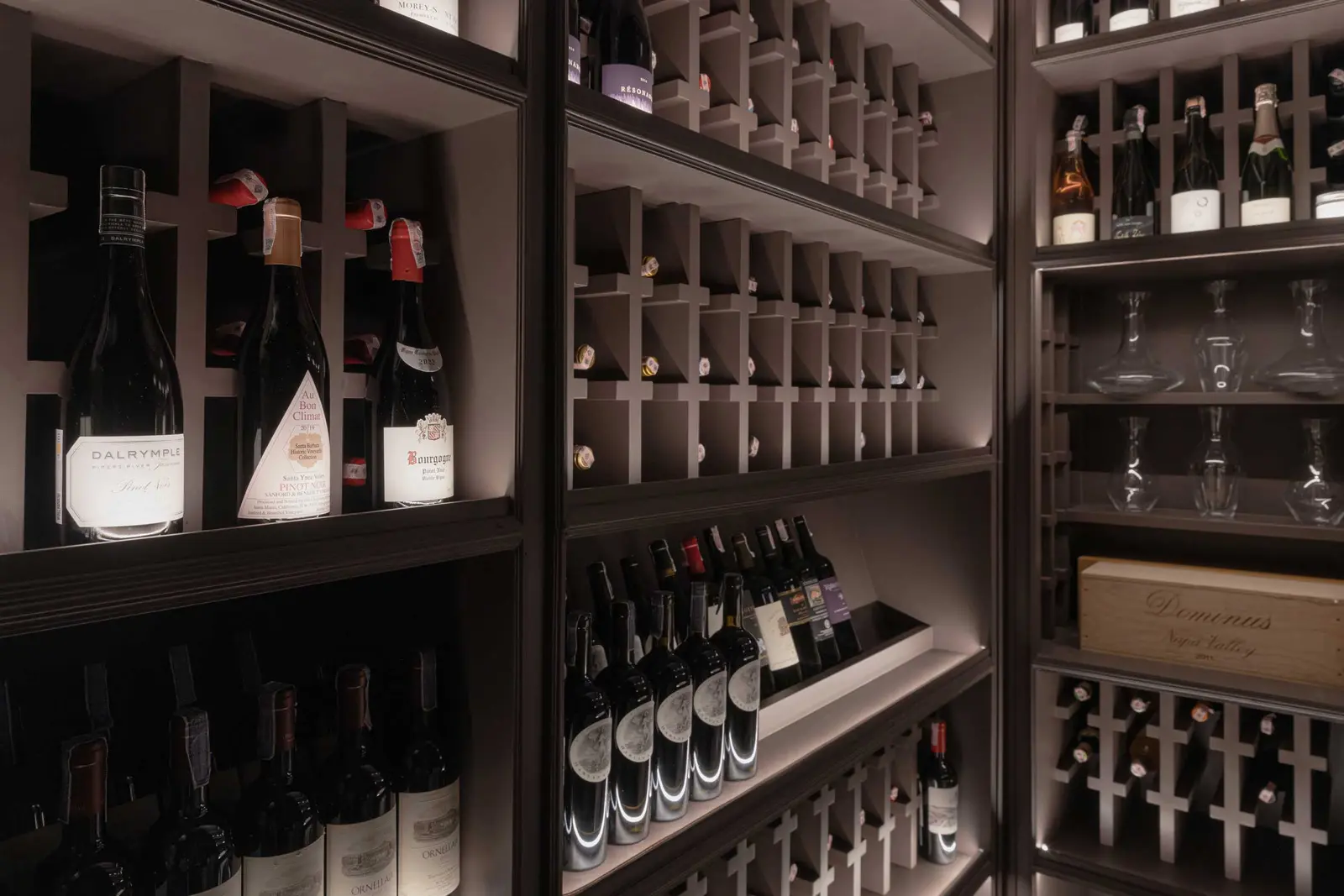 Backlit cellar racks filled with red and sparkling wine bottles, crystal decanters and a wooden case